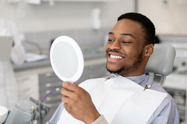 A young man with a beard smiles at the camera while holding a round mirror in front of his face, sitting in a dental chair with various dental equipment around him.
