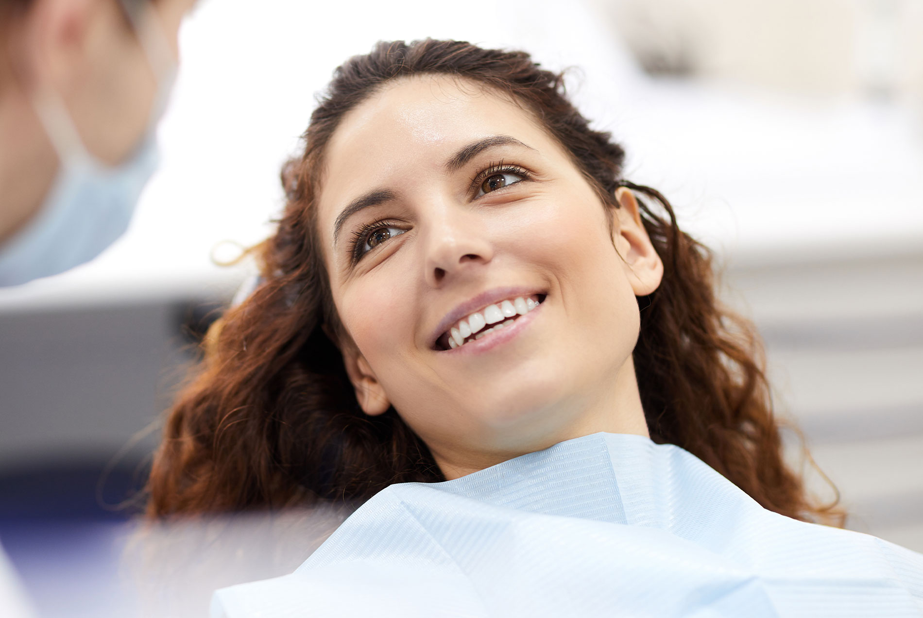 The image shows a woman seated in a dental chair with a smile on her face, looking at someone outside the frame, while a dental professional is attending to her, likely during a dental examination or procedure, with medical equipment visible in the background.