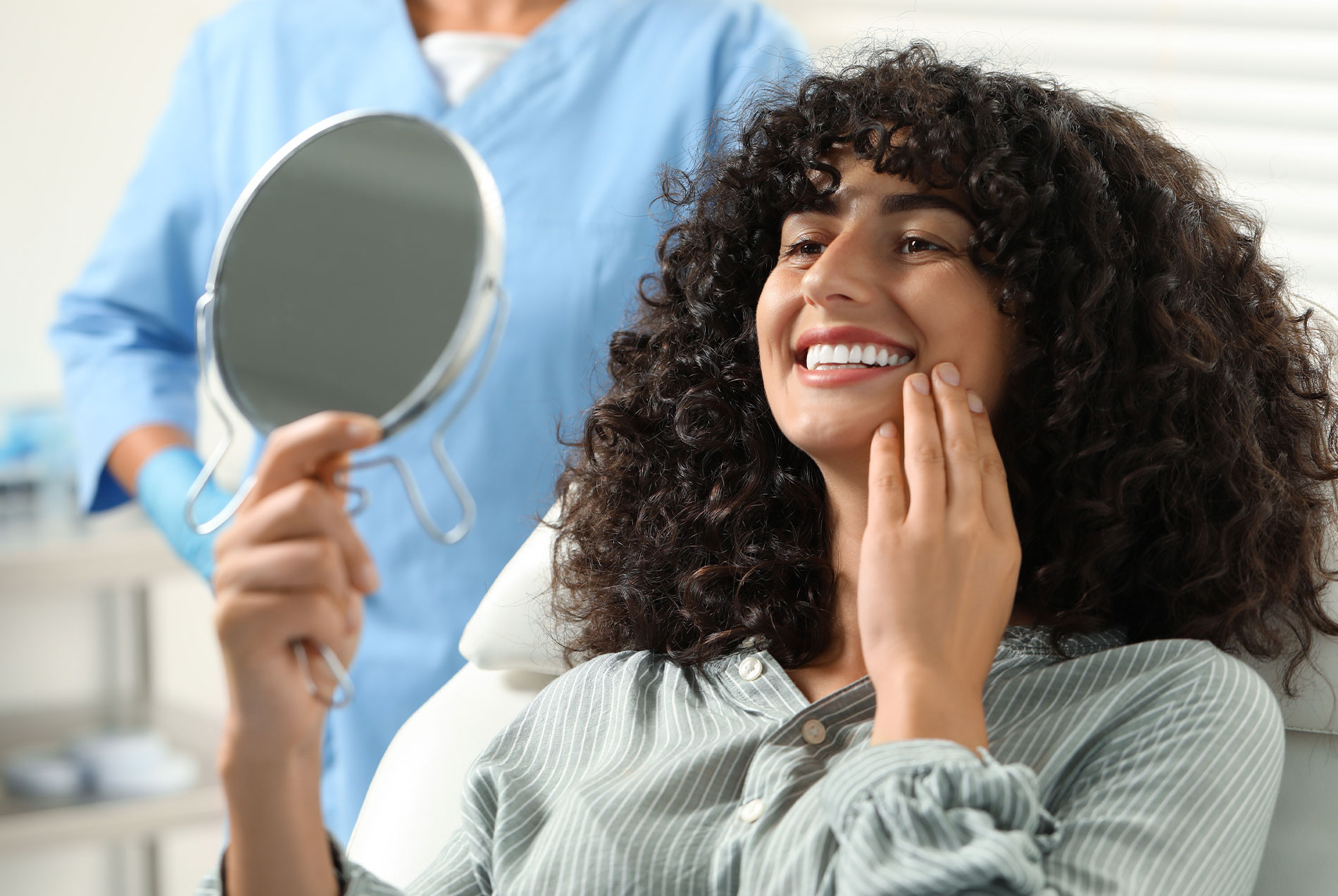 A person with curly hair sitting in a dental chair, smiling at the camera while holding a makeup mirror, with a dental hygienist standing behind them.