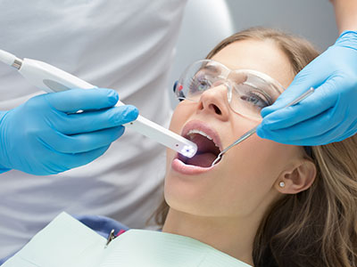 The image shows a dental professional using a device to clean a patient's teeth while the patient is seated in a dental chair, wearing protective eyewear and a surgical mask.